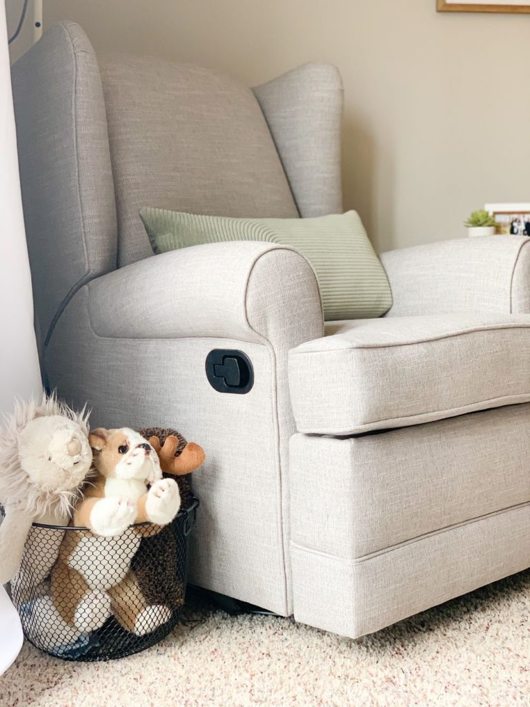 Pottery barn light grey, cream nursery chair with green pillow, white ikea lamp, white curtains and basket of stuffed animals in baby boy nursery.