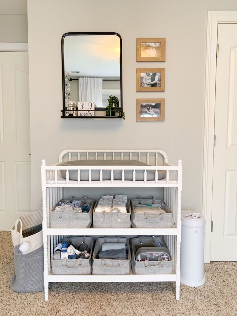 White Jenny Lind style changing table with neutral storage baskets with handles for easy access to changing essentials. Rod Iron mirror and wood framed photos, white changing pail and linen hamper.