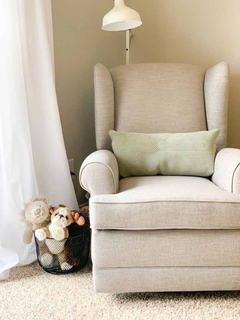 Pottery barn light grey, cream nursery chair with green pillow, white ikea lamp, white curtains and basket of stuffed animals in baby boy nursery.