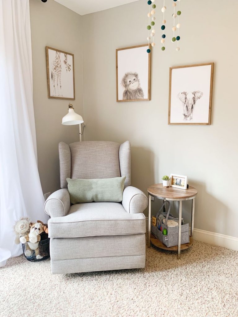 Pottery barn light grey, cream nursery chair with green pillow, white ikea lamp, white curtains and basket of stuffed animals in baby boy nursery. Neutral animal prints with white and wood details, and felt ball mobile.