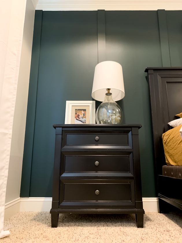 Black bedside table with glass lamp and white lampshade. Dark green board and batten wall behind it