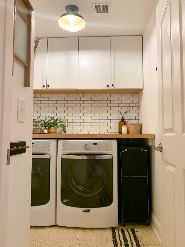 After modern farmhouse laundry room refresh with white cabinetry, white peel and stick subway tile with black grout, and natural butcher block. Jute rug, dark olive green laundry basket.