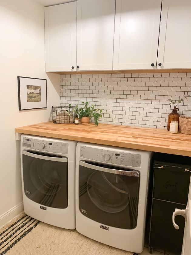 After modern farmhouse laundry room refresh with white cabinetry, white peel and stick subway tile with black grout, and natural butcher block. and laundry room oil print and jute rug