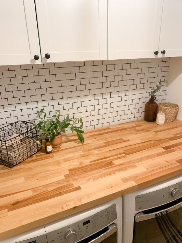 After modern farmhouse laundry room refresh with white cabinetry, white peel and stick subway tile with black grout, and natural butcher block.