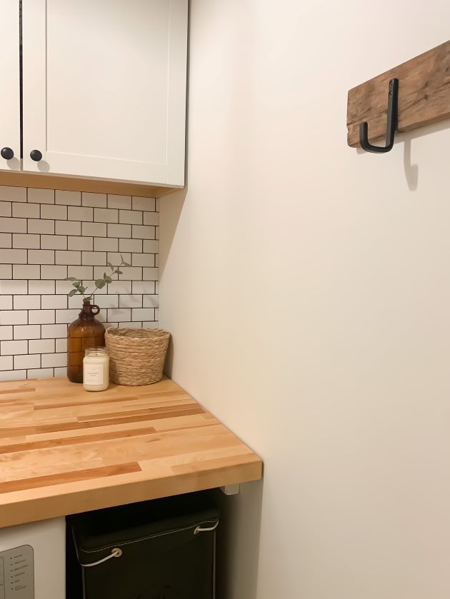 After modern farmhouse laundry room refresh with white cabinetry, white peel and stick subway tile with black grout, and natural butcher block. dark olive green laundry basket, reclaimed wood with modern black hook.
