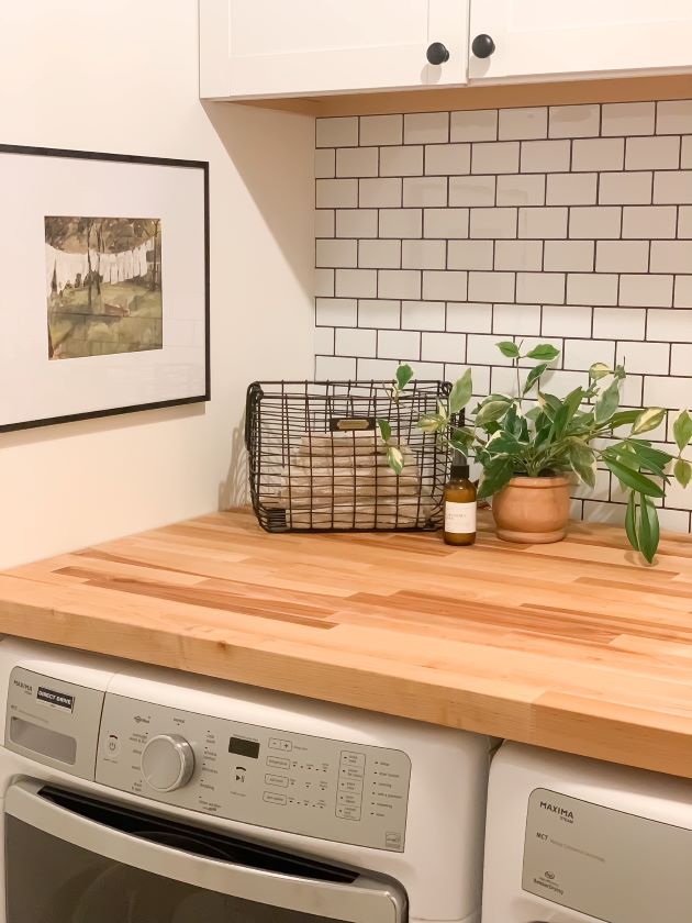 After modern farmhouse laundry room refresh with white cabinetry, white peel and stick subway tile with black grout, and natural butcher block. and laundry room oil print